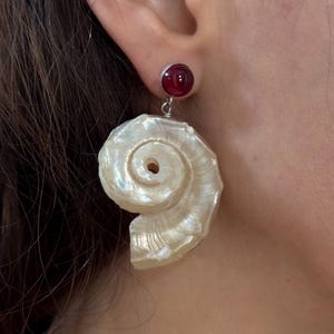 Op de afbeelding: Oorbellen met een rode edelsteenknop en een grote, parelmoer, spiraalvormige schelphanger. De schelphanger heeft een gedetailleerd, getextureerd oppervlak en een natuurlijke, organische vorm. Oorbellen voor doorboorde oren.