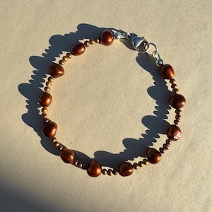 May include: A brown pearl bracelet with a silver clasp. The bracelet features large, irregularly shaped brown pearls interspersed with smaller, round, gold-coloured beads. The clasp is a simple, secure design.