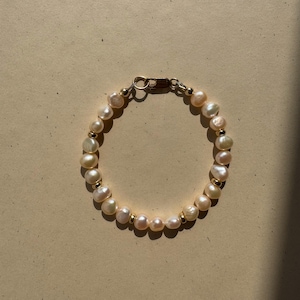 May include: A bracelet made of creamy-white, irregularly shaped pearls and small gold-coloured beads. The bracelet is fastened with a gold-toned clasp. The jewellery sits on a neutral-toned surface. A simple piece of jewellery.