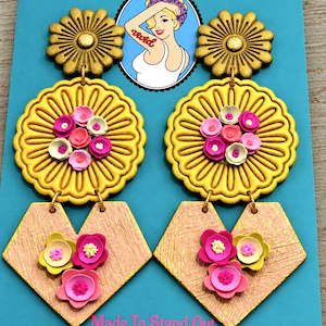May include: Dangle earrings featuring a gold-toned flower stud, a large yellow scalloped circular disc adorned with small pink and white flowers, and a textured peach-coloured heart-shaped pendant decorated with three small pink and yellow flowers.