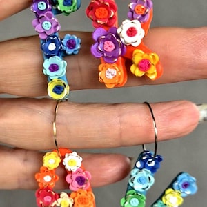 May include: Floral letter earrings in the letters A, R, and V. Each letter is decorated with small, colourful flowers in shades of blue, purple, orange, yellow, and white. The earrings are attached to gold-coloured hoops.