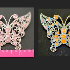 May include: A silver-toned butterfly brooch featuring intricate filigree metalwork and embedded round crystals. The left brooch displays light pink crystals, while the right brooch features orange crystals. A pink ruler shows the width is 2 inches.