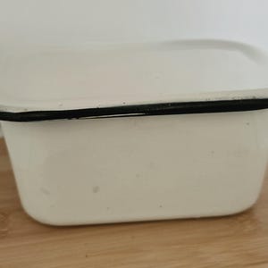 May include: A rectangular white enamel storage container with a matching lid. The container features rounded corners and a thin, dark black rim along the edge of both the lid and the base, providing a clean, high-contrast finish to the piece.