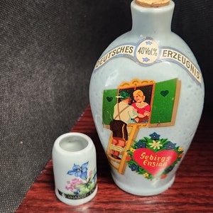 May include: A light blue ceramic bottle with a cork stopper and a small white ceramic cup. The bottle has a painted scene of two people and the words "DEUTSCHES ERZEUGNIS". The cup has a floral design.