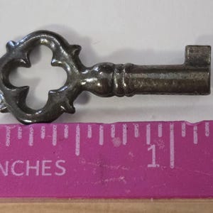May include: A decorative metal key with an ornate clover-shaped head. The key is silver-colored and measures approximately 1 inch in length, as indicated by the pink ruler in the image. The key has a vintage or antique aesthetic.