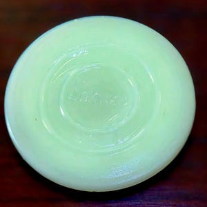 May include: A round, pale green glass object with a smooth, opaque finish. The center features a circular indentation with the embossed text "Fire-King" clearly visible. The piece has a slightly translucent quality and a uniform, light mint color.