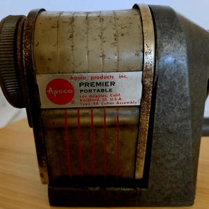 May include: A vintage Apsco Premier Portable pencil sharpener with a textured dark gray metal base and a light gray cylindrical body. The front label reads Apsco products inc. PREMIER PORTABLE Los Angeles, Calif. Rockford, Ill. U.S.A. Type 4A Cutter Assembly.