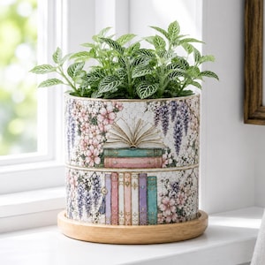 Pode incluir: Um vaso cilíndrico de cerâmica com uma estampa decorativa de livros empilhados, flores de cerejeira cor-de-rosa e flores de glicínia roxas. O vaso assenta num pires circular de madeira e contém uma planta verde com folhas nervuradas de branco.