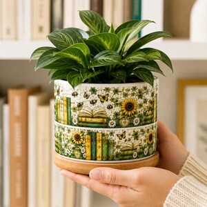May include: A cylindrical ceramic planter with a light-coloured base and a wooden saucer, featuring a repeating pattern of green books, yellow sunflowers, white daisies, butterflies, and bees, containing a lush green leafy plant.