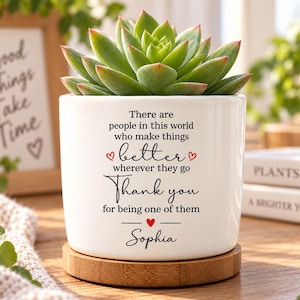 May include: A white ceramic cylindrical planter containing a green succulent plant, resting on a circular bamboo base. The planter features black printed text that reads: There are people in this world who make things better wherever they go Thank you for being one of them, with small red hearts and the name Sophia.