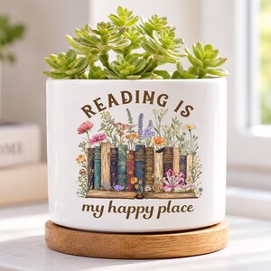Puede incluir: Una maceta cilíndrica de cerámica blanca con base de bambú que contiene una suculenta verde. La maceta presenta un gráfico de libros coloridos y flores silvestres con el texto marrón "READING IS my happy place" impreso en el frente.