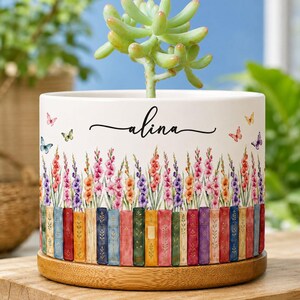 Op de afbeelding: Een witte cilindervormige keramische plantenpot met een houten voet, met een vetplant. De pot toont een kleurrijke illustratie van verticale boekruggen, bloemstengels in roze, paars en oranje, en vlinders, met de naam Alina erop gedrukt.