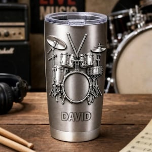 Op de afbeelding: Een zilverkleurige metalen beker met een verhoogd, reliëfontwerp van een drumstel met bekkens en drumstokken. De naam DAVID is in hoofdletters bij de basis gegraveerd. De beker heeft een doorzichtig plastic deksel en een geborstelde afwerking.