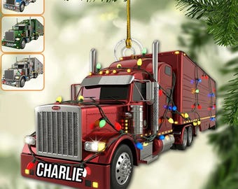 May include: A red semi-truck Christmas ornament with the name "CHARLIE" on the front. The truck is decorated with colorful Christmas lights and has a trailer attached. The ornament is hanging from a gold string.