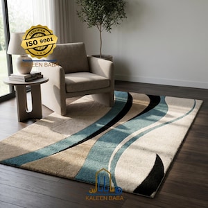 Op de afbeelding: Een rechthoekig vloerkleed met een beige achtergrond en een vloeiend ontwerp in teal, zwart en bruin. Het vloerkleed staat in een woonkamer met een stoel, een bijzettafel en een lamp. Het vloerkleed heeft de tekst "KALEEN BABA" en "ISO 9001".