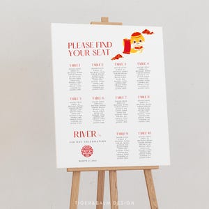 May include: A white seating chart on an easel with red text that reads "PLEASE FIND YOUR SEAT". The chart lists names under table numbers 1-10. A red and gold illustration is in the upper right corner. The text "RIVER'S 100 DAY CELEBRATION" is also present.