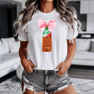 May include: White t-shirt with a pink bow and a bottle of Tajín seasoning. The bottle is green, red, and white with the word "Tajín" printed on it.
