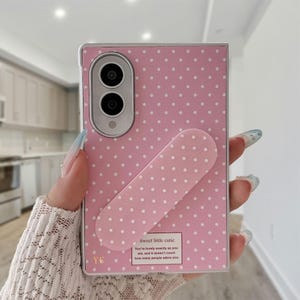 May include: A rectangular pink phone case with a white polka dot pattern. The case features a vertical camera cutout, a matching pink pill-shaped phone grip, and a small rectangular label with the text Sweet little cutie. You're lovely exactly as you are, and it doesn't count how many people adore you.