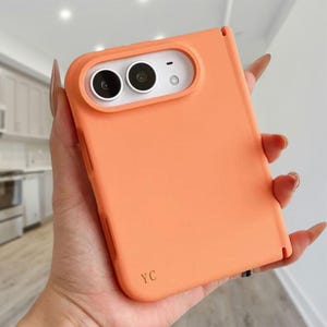 May include: A solid matte orange phone case for a foldable smartphone, featuring a raised white camera housing with two circular lenses and a small flash. The lower corner of the case is embossed with the letters YC in a simple serif font.