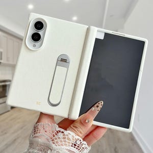 Könnte beinhalten: Eine weiße, faltbare Smartphone-Hülle mit strukturierter Oberfläche, ausgestattet mit einem Ausschnitt für zwei Kameras, einem silbernen Metallständer auf der Rückseite und den goldenen Buchstaben YC. Die Hülle ist offen und zeigt einen schwarzen Rahmen.
