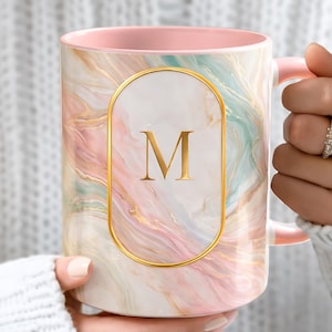 May include: A ceramic mug with a pink interior and handle, featuring a marble design in shades of pink, blue, and gold. A gold-framed oval on the front displays the letter "M". The mug is held by a person.