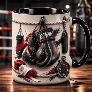 Könnte beinhalten: Weißer Keramikbecher mit schwarzem Innenraum und Griff, mit Box-Motiv. Das Design umfasst schwarze Boxhandschuhe mit "ETHAN", eine Stoppuhr und rote und weiße Bandagen. Der Becher ist mit einem Seilmotiv verziert.