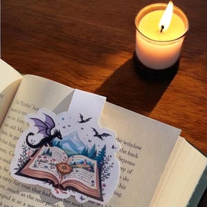 May include: A purple and black dragon bookmark with a book, mountains, and bats in the background. The bookmark is on a book with the text "The Plinths' lux" visible.