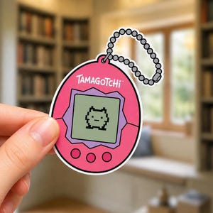 May include: A pink Tamagotchi sticker featuring a digital screen with a pixelated character, three circular buttons below the screen, and a silver ball chain attachment at the top. The sticker has a white border and the word Tamagotchi printed.