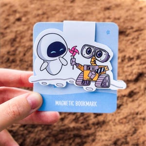 May include: A magnetic bookmark featuring the characters Eve and Wall-E from the animated film. Eve is white with a black oval face, and Wall-E is yellow with large, round eyes. The bookmark is light blue and has the words "MAGNETIC BOOKMARK".