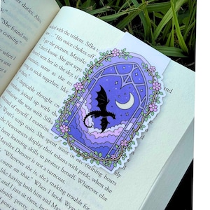 May include: A rectangular bookmark featuring a purple arched window frame decorated with pink flowers and green vines. Inside the window, a black dragon silhouette flies against a night sky with a white crescent moon and purple clouds.
