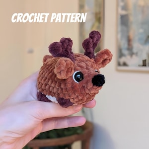May include: A crocheted deer with brown and burgundy yarn. The deer has large black eyes and a black nose. The text "CROCHET PATTERN" is in white letters at the top of the image. The deer is held in a person's hand.