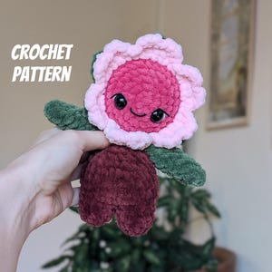 May include: A handmade crochet flower plushie with a smiling face. The flower has a pink and red center, pink petals, green leaves, and a burgundy base. The text "CROCHET PATTERN" is in the upper left corner.