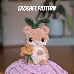 Könnte beinhalten: Häkelanleitung für einen braunen und rosafarbenen Hamster mit einem grünen, blättrigen Stiel auf dem Kopf und einer gelben Blume in der Pfote. Der Hamster sitzt auf einem rosa Wollknäuel. "CROCHET PATTERN" steht in weißen Buchstaben auf schwarzem Hintergrund.