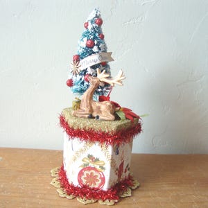 May include: A hexagonal decorative box featuring a vintage-style Christmas print with red ornaments and candles, trimmed with red tinsel. Atop the box sits a miniature flocked green Christmas tree with red berries and a deer figurine with antlers.
