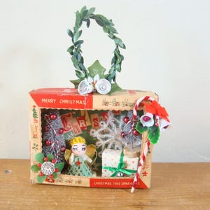 May include: A rectangular cardboard box labeled MERRY CHRISTMAS containing a miniature angel figurine, two silver bottle brush trees, a white snowflake, and a gift box. The top features a green leaf wreath with two white bells and a candy cane.
