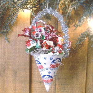 May include: A festive Christmas ornament shaped like a cone, filled with holiday decorations. The cone is made from a Sno-Kone paper cup, adorned with snowflakes and the Sno-Kone logo. The ornament features a silver tinsel handle and is filled with red and white embellishments.