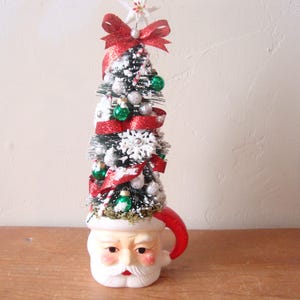 May include: A whimsical Christmas tree decoration. The tree is atop a Santa Claus mug, with a red handle. The tree is decorated with silver ornaments, green baubles, red ribbon, and a white star on top. The mug has a jolly face.