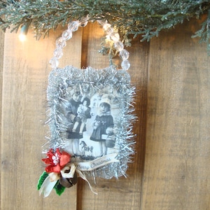 May include: A vintage Christmas ornament featuring a black and white photograph of children, framed with silver tinsel. The ornament is adorned with a red poinsettia, a bell, and a banner that reads "Merry Christmas". It hangs from a beaded string.