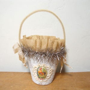 May include: A handmade white paper mache basket with a decorative rooster image. The basket has a beige handle, a ruffled top, and silver tinsel trim. A decorative ribbon is tied on the side. The basket is on a wooden surface.