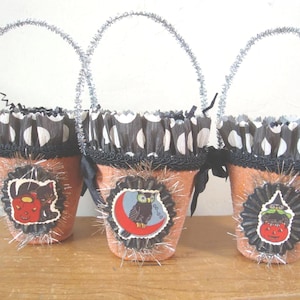 May include: Three orange glittery pots with silver tinsel handles and black and white polka dot paper ruffles. Each pot features a central circular medallion with Halloween-themed illustrations of a black cat, an owl, and a witch.