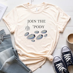 May include: Cream-colored t-shirt with the text "JOIN THE 'PODY" and a disco ball graphic. Below the text are several blue and gray pill bug illustrations. The shirt is paired with blue jeans and navy blue sneakers.
