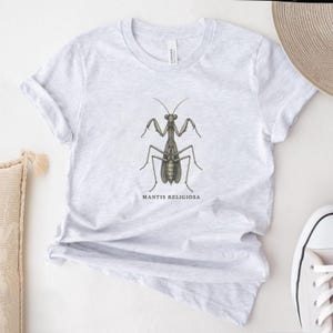 May include: Light gray t-shirt featuring a detailed illustration of a praying mantis. The insect is depicted in shades of brown and green, with the scientific name "MANTIS RELIGIOSA" printed below. The shirt has short sleeves and a crew neck.