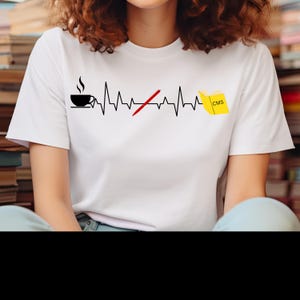 May include: White t-shirt with a black graphic of a coffee cup, a heartbeat line, a red X over a pen, and a yellow book with "CMS" on it. The shirt is a crew neck style.