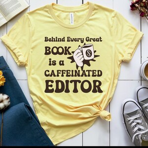 May include: A light yellow t-shirt with the text "Behind Every Great Book is a Caffeinated Editor" in brown lettering. The graphic includes a hand holding a coffee mug with a dollar sign. The shirt is knotted at the bottom.