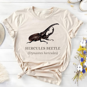May include: Beige t-shirt featuring a detailed illustration of a Hercules beetle, with the text "HERCULES BEETLE (Dynastes hercules)". The beetle is depicted in shades of brown and black, with prominent horns. The shirt is on a white wooden surface.
