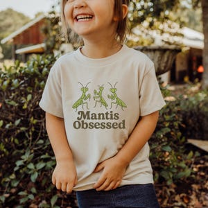 May include: Beige short-sleeved t-shirt featuring three green praying mantis illustrations above the text "Mantis Obsessed" in brown. The shirt is a neutral color, suitable for various outfits.