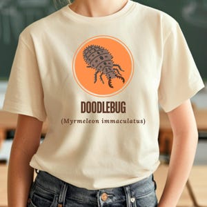 May include: Cream-colored t-shirt featuring a cartoon doodlebug illustration inside an orange circle. The word "DOODLEBUG" is printed below the graphic, with the scientific name (Myrmeleon immaculatus) underneath.