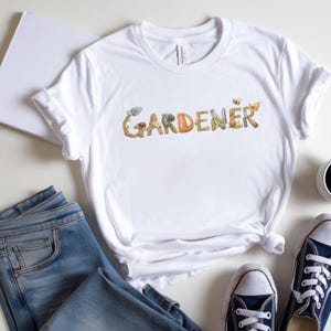 Peut inclure: T-shirt blanc avec le mot "GARDENER" en lettres colorées sur le thème de la nature. Le design comprend des éléments tels qu'une pousse, un escargot et un papillon. Le t-shirt est présenté avec un jean bleu et des baskets bleues.