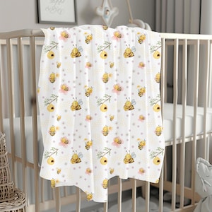 May include: A white baby blanket with a repeating pattern of yellow bees, pink flowers, and honeycomb. The bees are cartoon-like and have closed eyes.