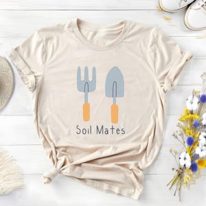 Peut inclure: Un t-shirt crème avec un motif de fourche et de truelle de jardin avec des manches orange et le texte "Soil Mates". Le t-shirt est plié, avec des manches retroussées, suggérant un style décontracté. Le design est simple et charmant.